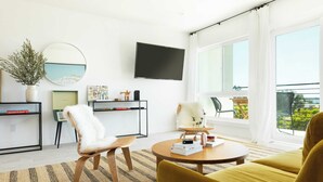 A modern living room with a sofa, armchair, and coffee table, a flat-screen TV mounted on the wall, and a large window offering a view of the outdoors. - Avalon I by AvantStay Contemporary San Diego Condo w Large Balcony (San Diego)
