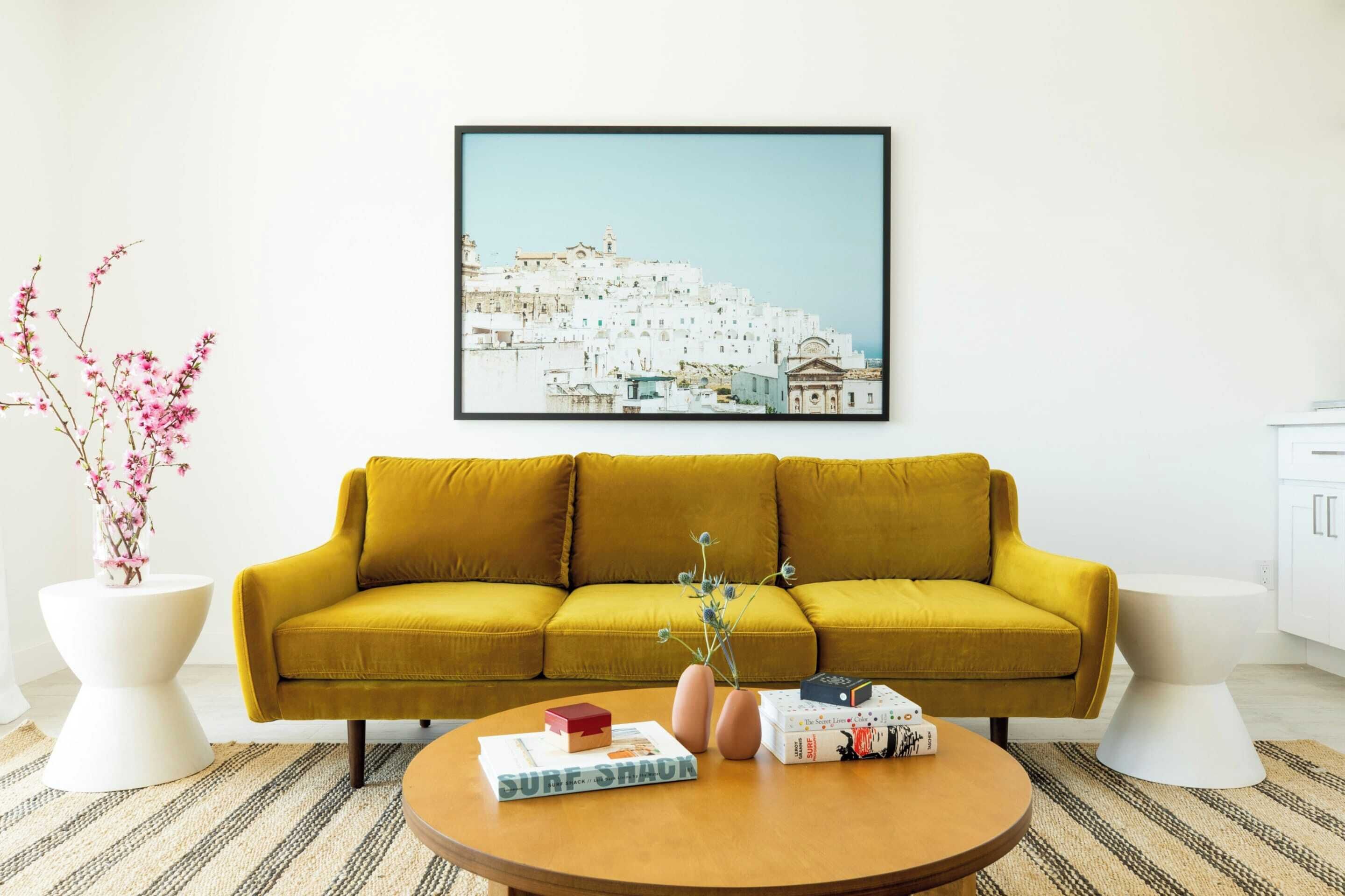 Een woonkamer met een mosterdgele bank, een ronde salontafel met het boek 'Surf Shack' en een lijst met een foto van een kustplaatsje aan de muur.