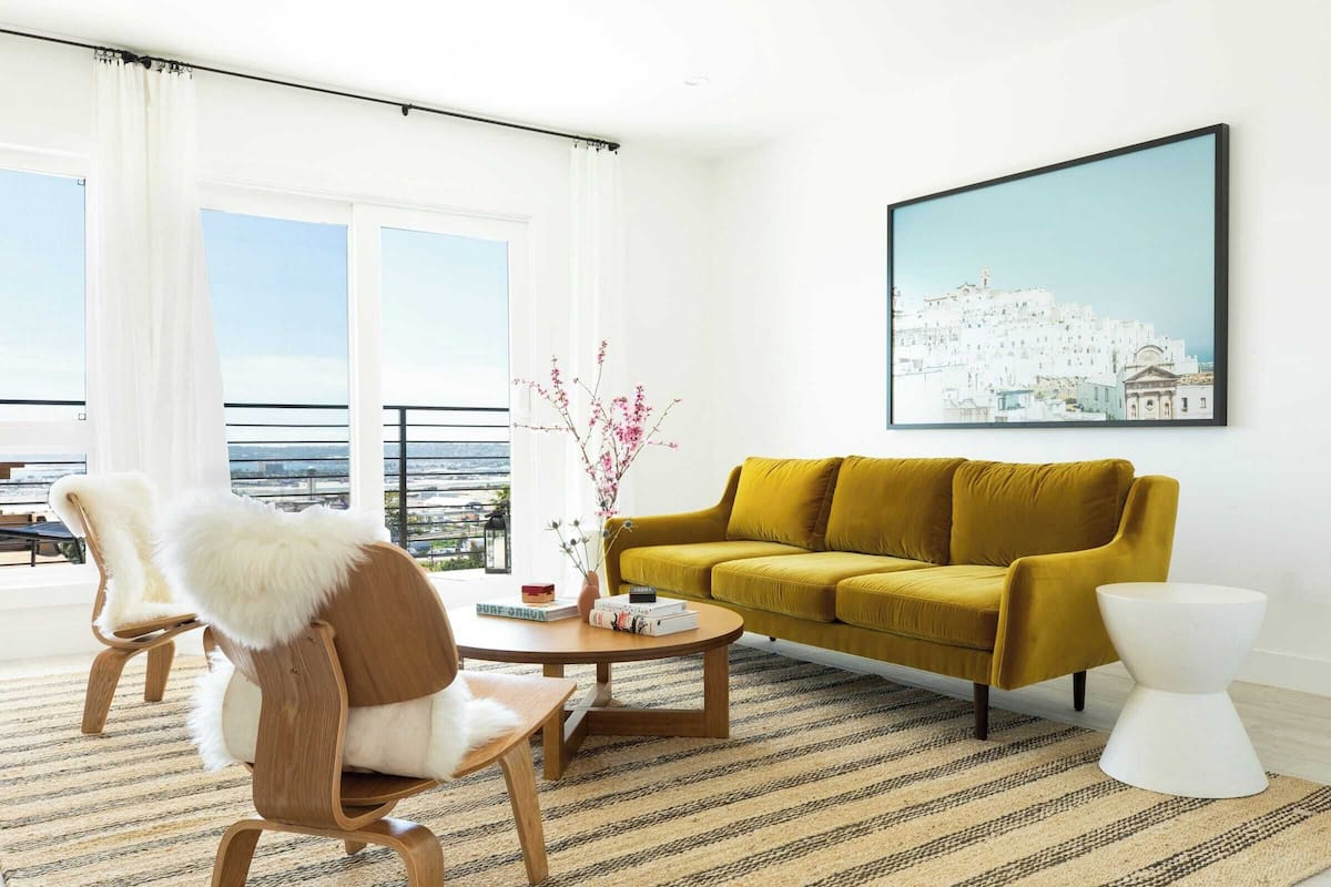 A modern living room with a yellow sofa, wooden chairs, a coffee table, and a framed cityscape picture on the wall.