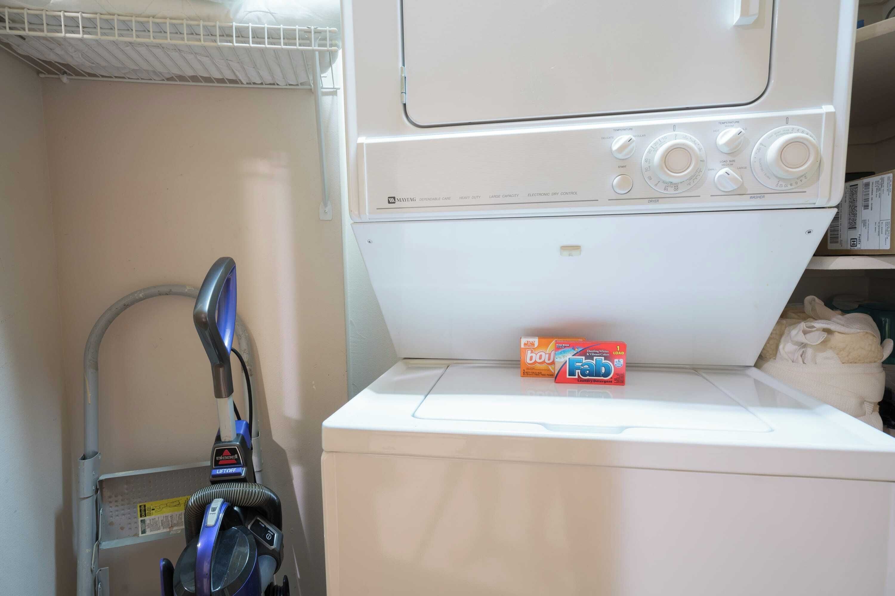 A front-loading washing machine with a control panel, a vacuum cleaner, and a box of laundry detergent.