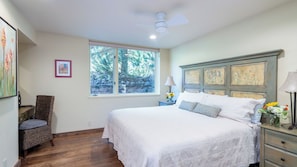 Un dormitorio con una cama grande, una ventana con vistas a árboles, un ventilador de techo y una mesita de noche con una lámpara.