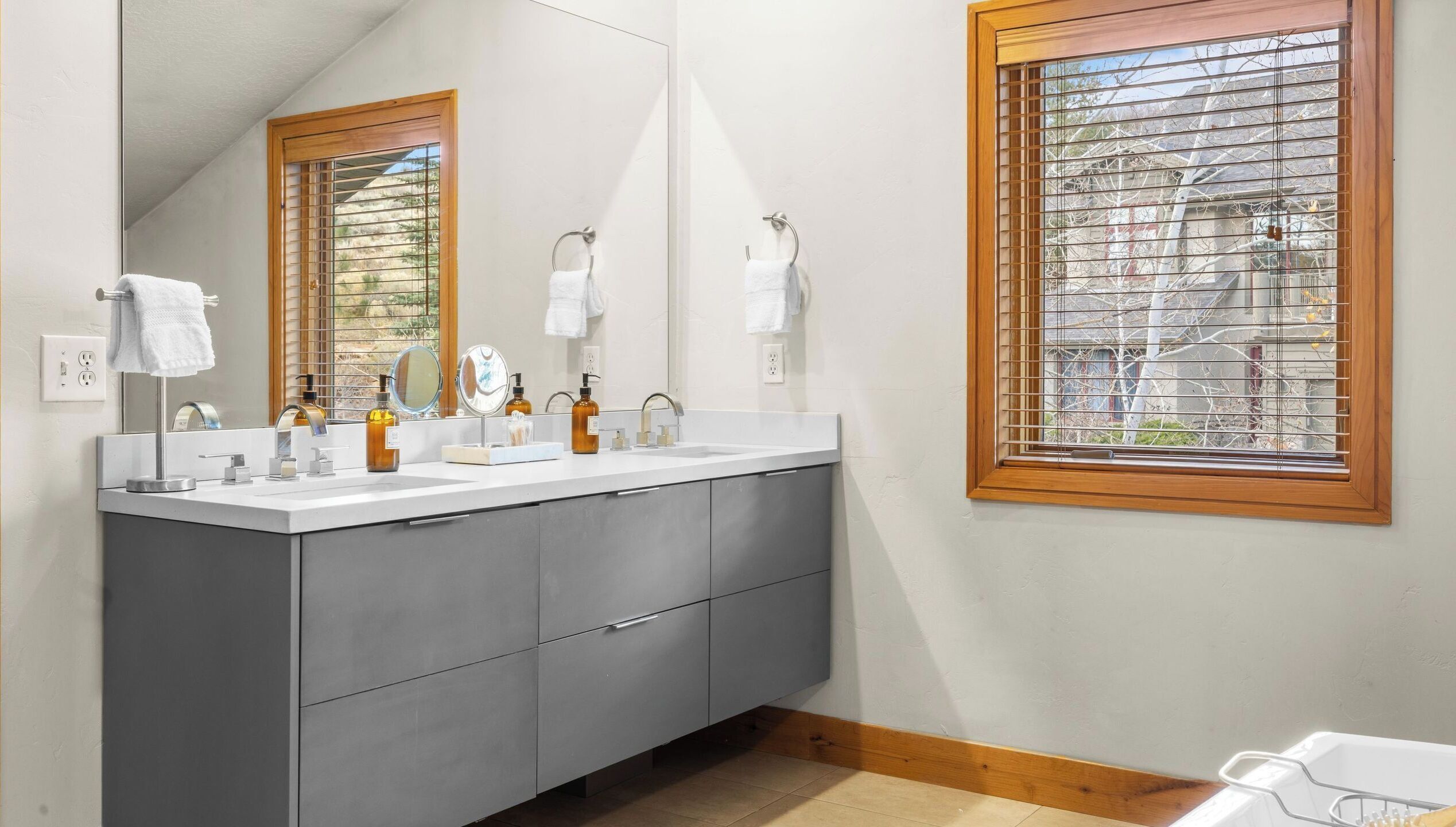 A modern bathroom with a large mirror, two sinks, and a window with blinds.
