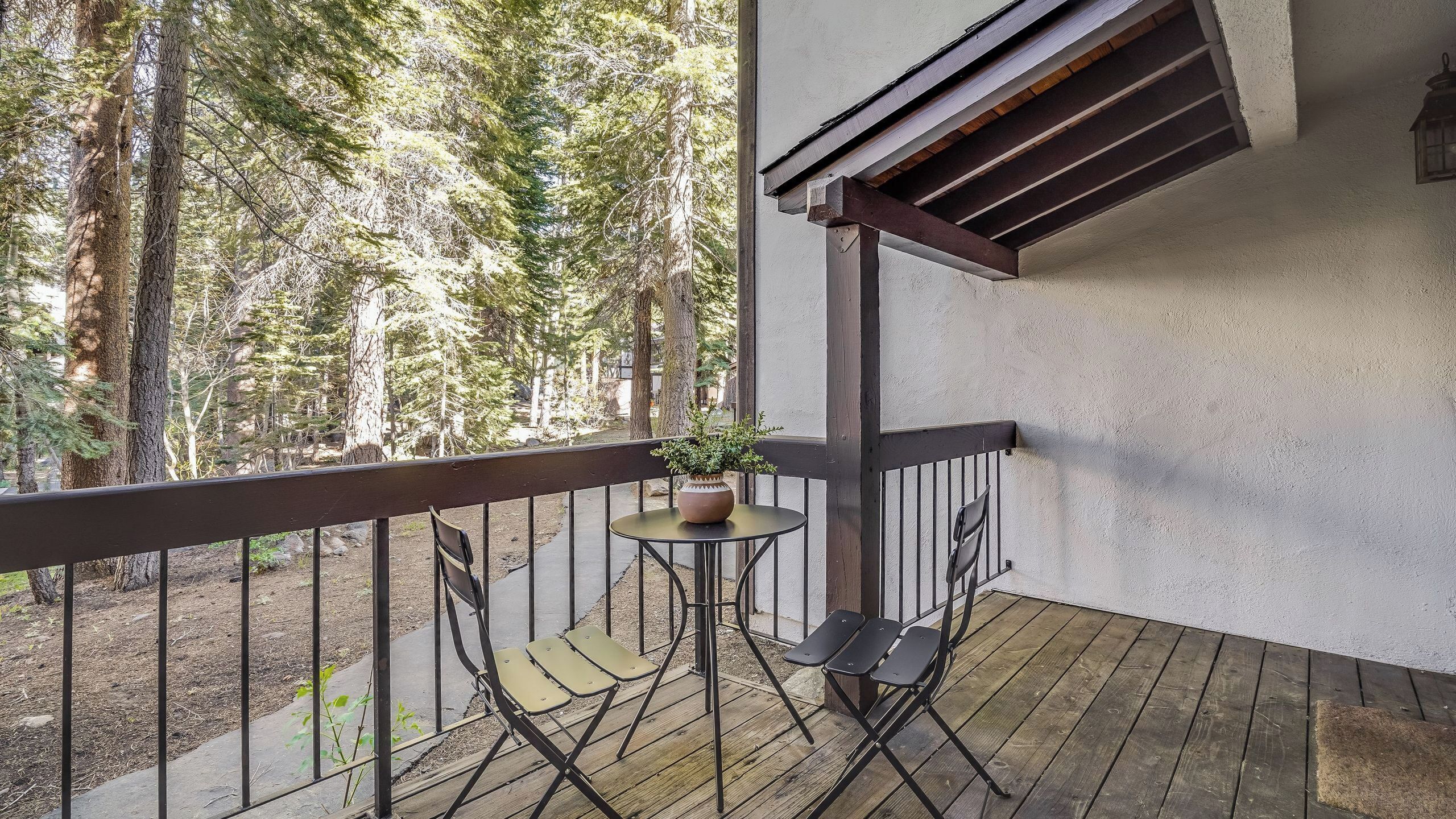 Un balcon avec une petite table et deux chaises, un pot de fleurs et une vue sur une forêt.