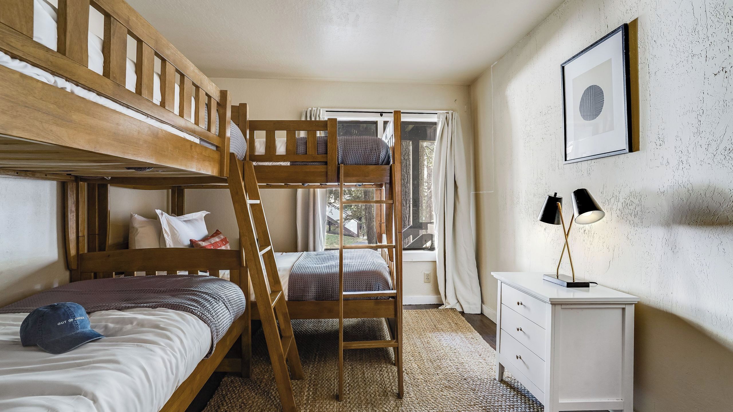 A bunk bed room with a ladder, a desk, a lamp, and a window with a view of trees.
