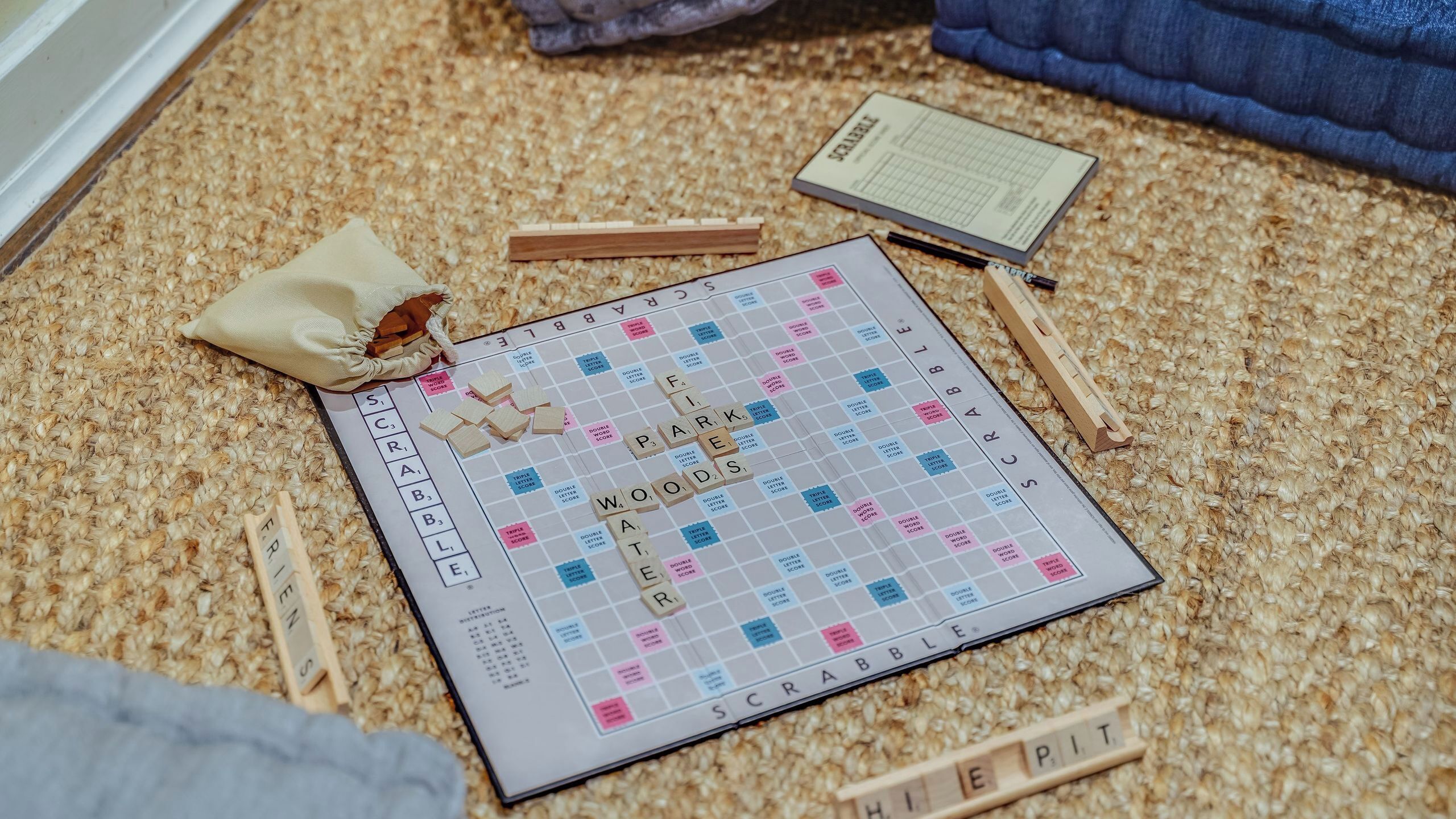 En Scrabble-spel på ett mattbeklätt golv, med spelbrickor, en påse och en bok.