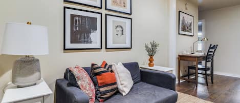 A living room with a dark grey sofa, a white side table, a lamp, and framed artwork on the wall.