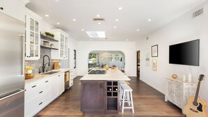 A modern kitchen with white cabinetry, a central island, and a dining area with a wooden table. - Vista Del Mar by Avantstay Stunning Spanish Inspired Home w/ Pool, Hot Tub & Rooftop Patio (La Jolla)