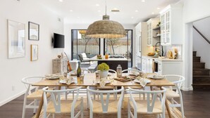 A dining area with a wooden table, white chairs, and a hanging light fixture. - Vista Del Mar by Avantstay Stunning Spanish Inspired Home w/ Pool, Hot Tub & Rooftop Patio (La Jolla)