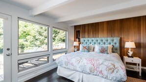A bedroom with a large bed, wooden paneling, two bedside tables with lamps, and a view of greenery through large windows. - Rancho del Mar by Avantstay One Block From Beach (Arch Cape)
