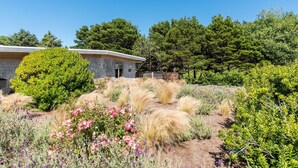 A house with a stone exterior, surrounded by a garden with various plants and a large tree. - Rancho del Mar by Avantstay One Block From Beach (Arch Cape)