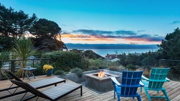 A wooden deck with lounge chairs, a fire pit, and a view of the ocean at sunset.