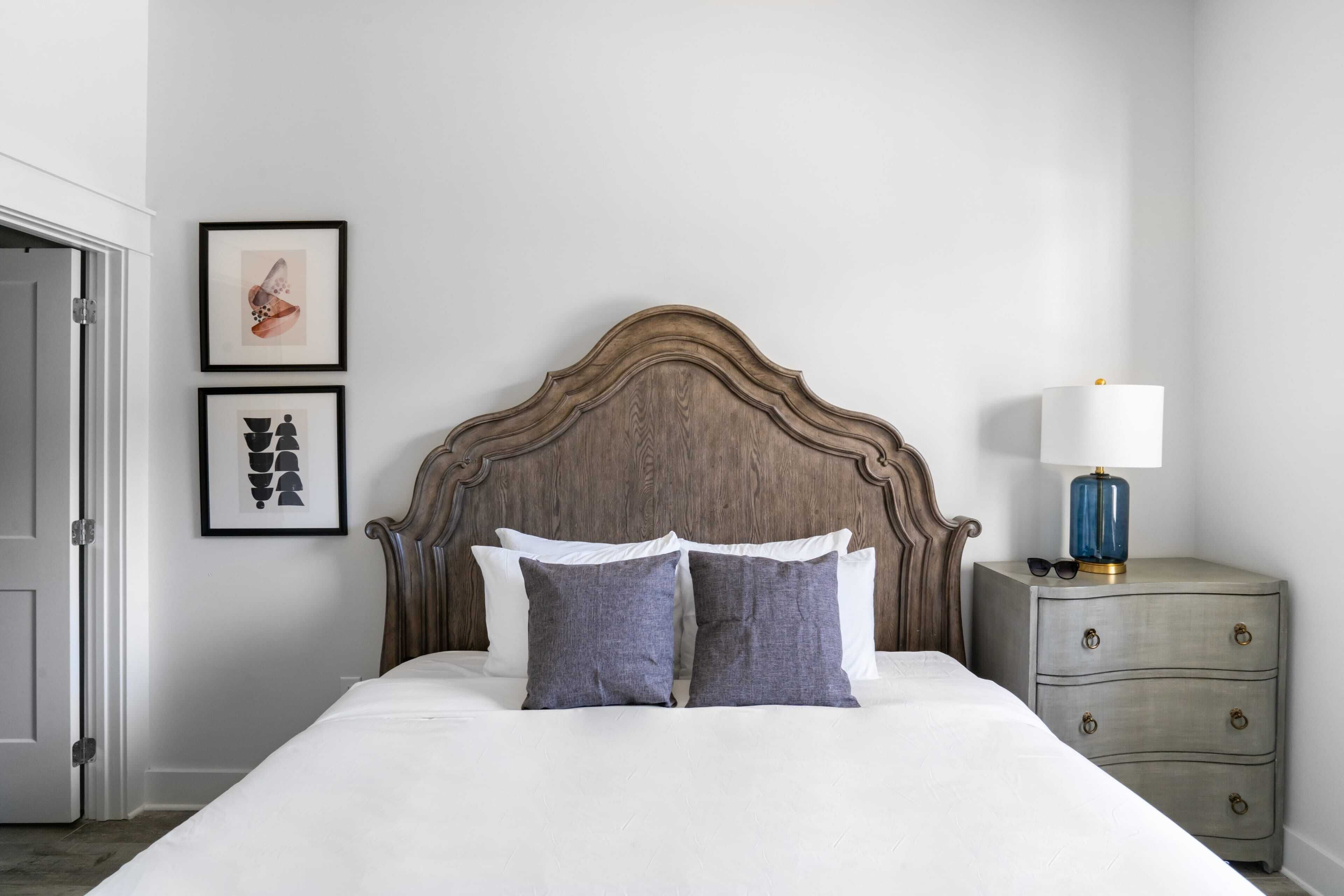 Un lit bien fait, avec une tête de lit en bois, une table de chevet avec une lampe et deux tableaux encadrés accrochés au mur.