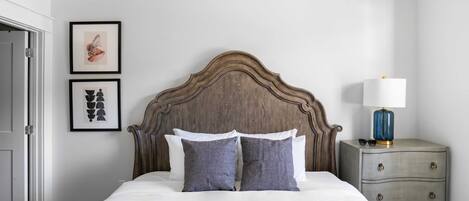 Un lit bien fait, avec une tête de lit en bois, une table de chevet avec une lampe et deux tableaux encadrés accrochés au mur.