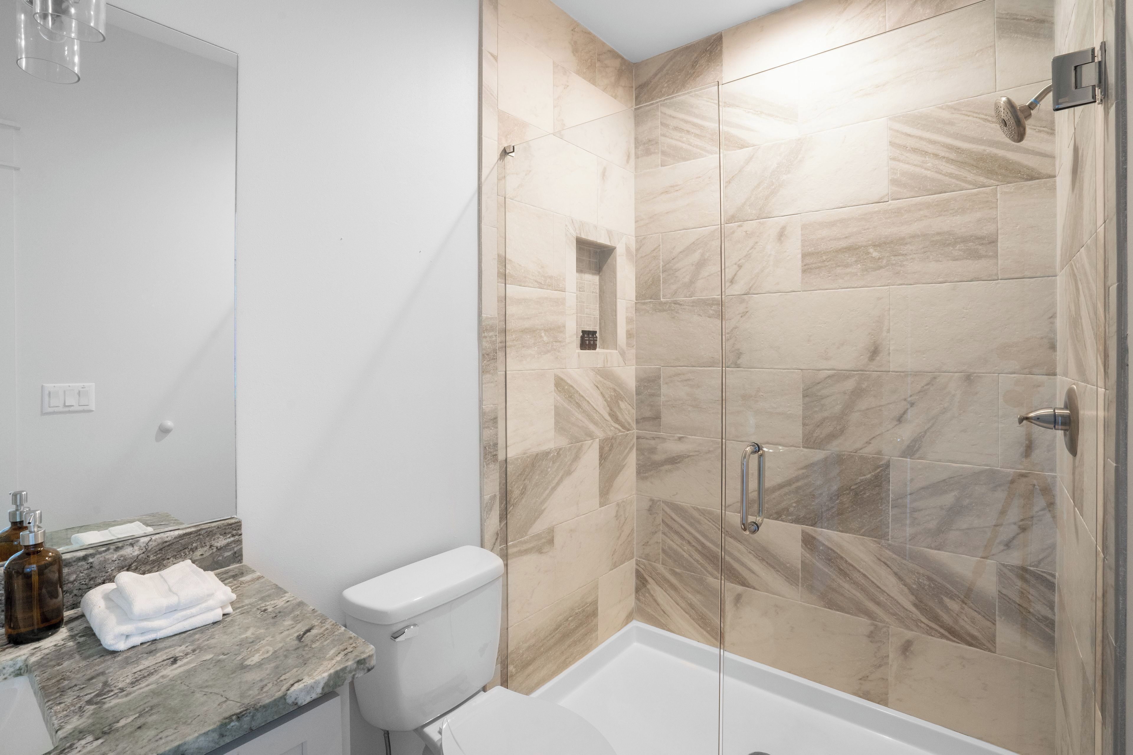 A bathroom with a marble countertop, a toilet, a shower with glass doors, and a small shelf.