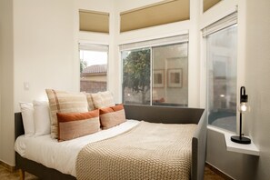 A bedroom with a bed, pillows, a bedside table, and a window with blinds. - Petit Sahara by Avantstay Desert Escape w/ Pool, Outdoor Dining & Table Tennis! (Indio)