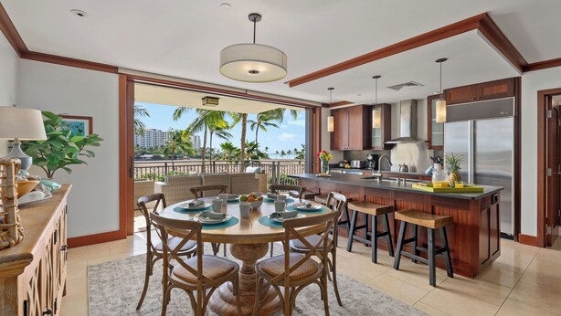 A modern kitchen with a central island, dining area with chairs, and a view of palm trees and buildings. - Hale Moana by Avantstay Incredible Hawaii Beach Resort w/ Pool & Hot Tub! (Kapolei)