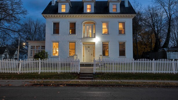 Une maison blanche à deux étages, avec une clôture en piquets blancs, une entrée centrale avec le numéro 94 sur la porte, et de nombreuses fenêtres.