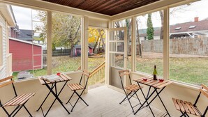 Une véranda couverte avec des chaises et des tables en bois, de grandes fenêtres et une vue sur le jardin arrière.