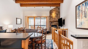 A cozy interior with a stone fireplace, wooden ceiling beams, a dining area with a table and chairs, and a large window overlooking a snowy landscape. - Riverside Condos C202 by Avantstay Condo Close To Downtown, Town Park & Ski Lift #8 (Telluride)