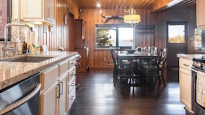 A spacious kitchen with wooden walls, a granite countertop, and a dining area with a table and chairs. - Shoreline Lakehouse by Avantstay Majestic Lakefront Cabin w/ Hot Tub & Fire Pit! (Big Bear Lake)