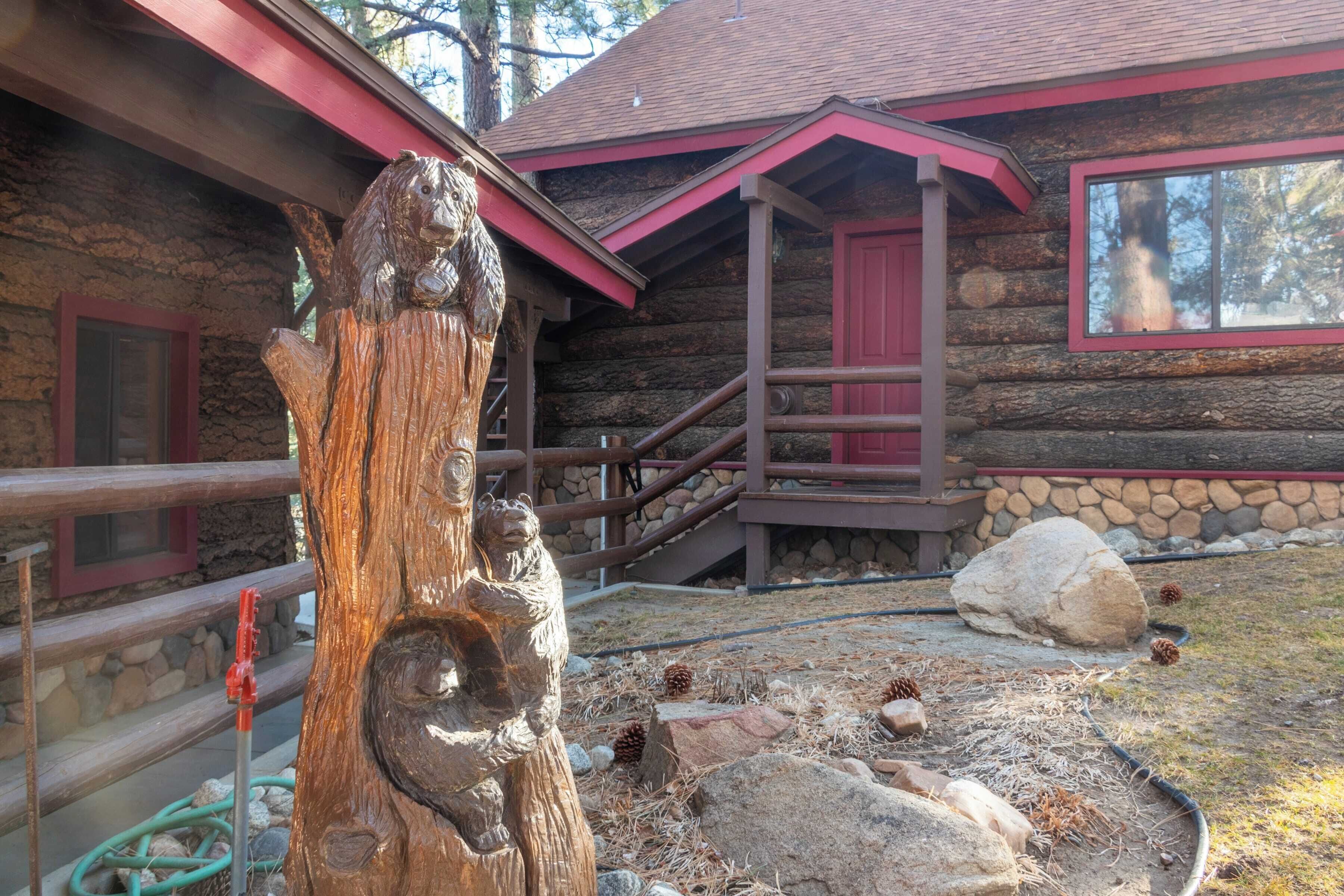 A rustic log cabin with a wooden sculpture in the foreground, a stone staircase, and a window with a red frame.