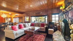 L’intérieur chaleureux d’un chalet avec un plafond en bois, une cheminée en pierre, un canapé avec des coussins rouges et un coin repas doté de meubles en bois.