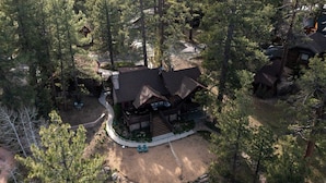 A cabin surrounded by dense forest, with a dirt driveway leading to a wooden deck and outdoor seating area. - Shoreline Lakehouse by Avantstay Majestic Lakefront Cabin w/ Hot Tub & Fire Pit! (Big Bear Lake)