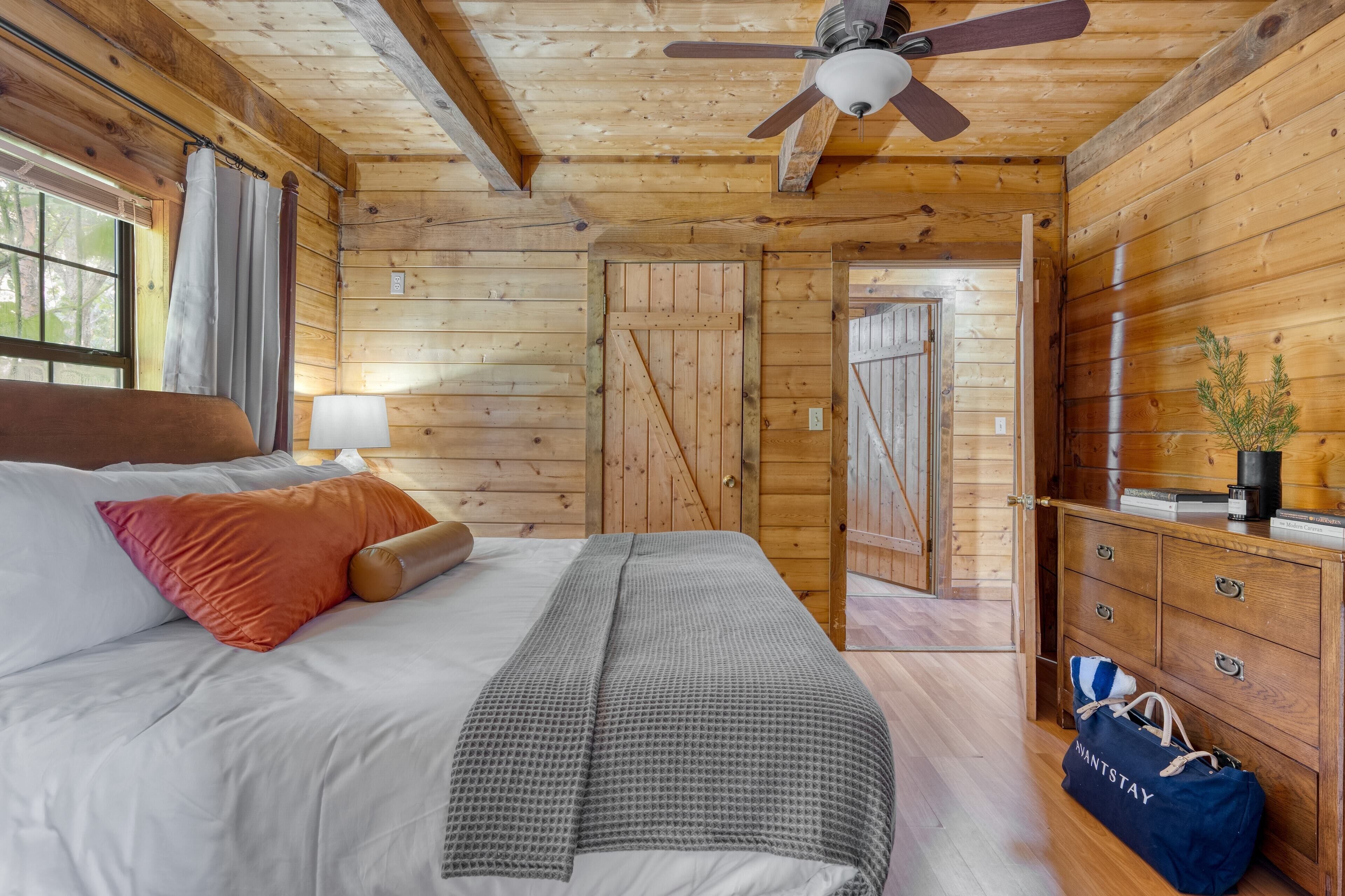 A wooden cabin bedroom with a bed, a ceiling fan, a dresser, and a door leading to another room.