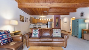 A living room with a brown leather sofa, a wooden ceiling, and a kitchen in the background. - Riverside Condos D01 by Avantstay Condo Close To Downtown, Town Park & Ski Lift #8 (Telluride)