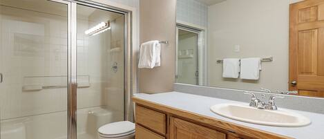 A bathroom with a glass shower enclosure, a toilet, a sink with a mirror, and wooden cabinets.