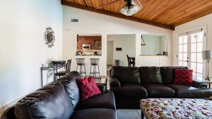 A living room with a brown leather sofa, a coffee table, and a patterned ottoman. There is a ceiling fan, a microwave, and a kitchen area visible in the background. - Desert Ridge by Avantstay Spacious Oasis w/ Pool & Hot Tub (Scottsdale)