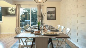 A dining area with a long table set for four, surrounded by chairs. A chandelier hangs above, and a window provides a view of the outdoors.