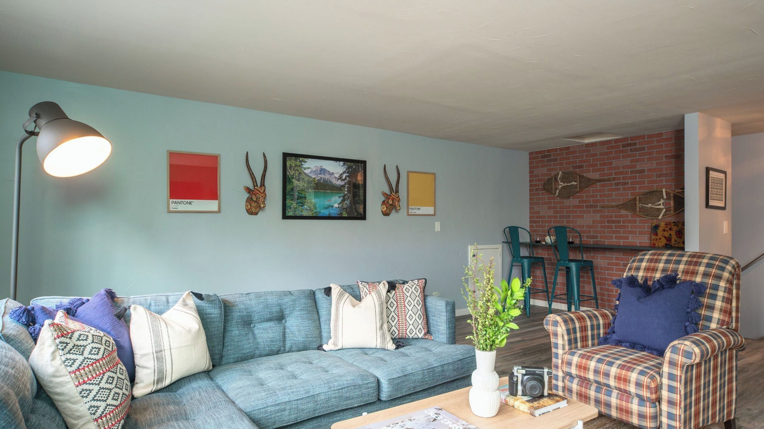 A living room with a blue sofa, a plaid armchair, a coffee table with a camera and books, and a wall with framed pictures and a lamp.