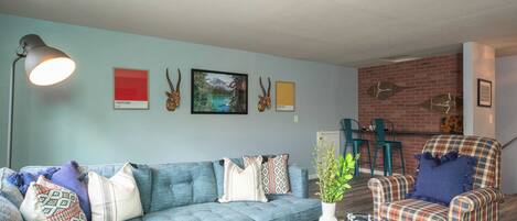 A living room with a blue sofa, patterned cushions, a coffee table with a magazine, a lamp, and wall art including animal horns and a mountain landscape.