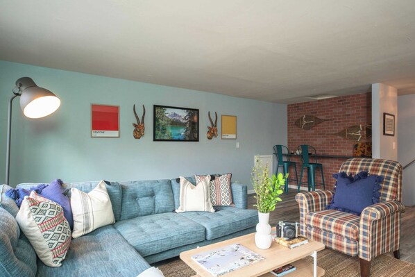 A living room with a blue sofa, patterned cushions, a coffee table with a magazine, a lamp, and wall art including animal horns and a mountain landscape.