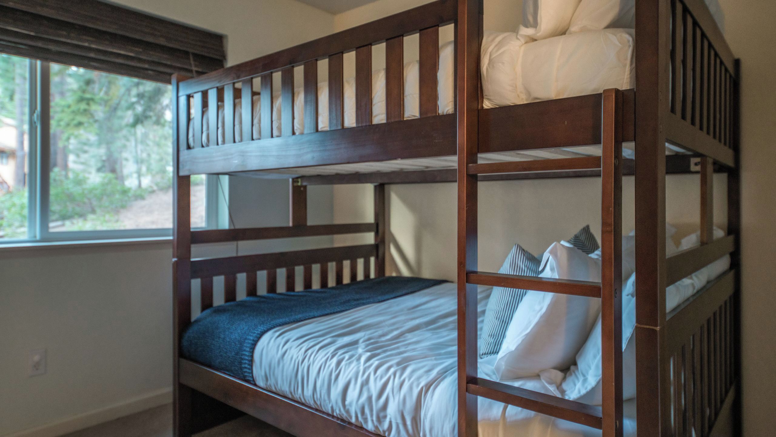 A bunk bed with a window view, a bedside table, and a bed with pillows and a blanket.