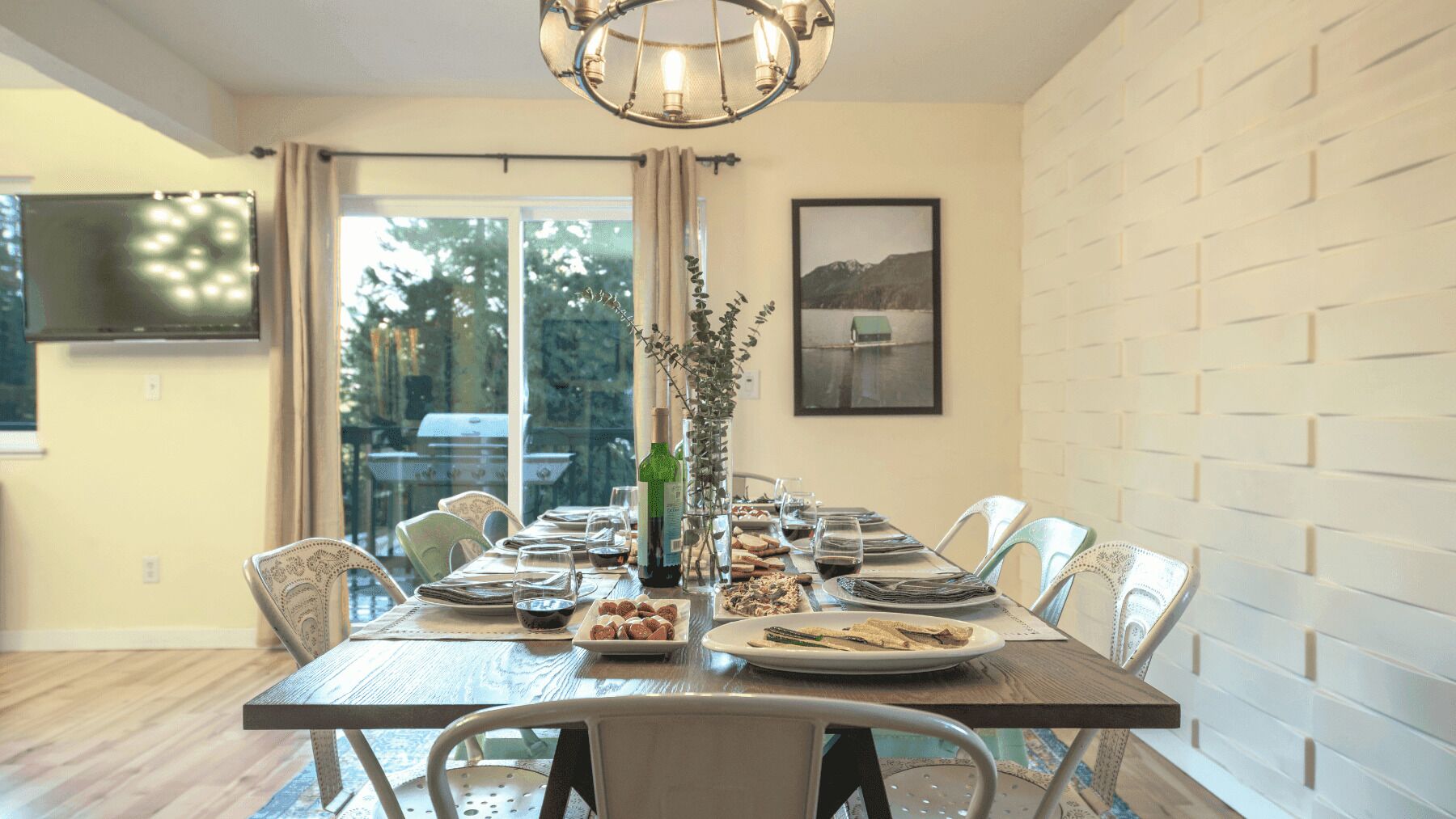 Une salle à manger avec une grande table dressée pour six personnes, un lustre, un téléviseur fixé au mur et une fenêtre donnant sur des arbres.