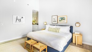 A bedroom with a bed, bedside tables, a nightstand, a small stool, and framed pictures on the wall.