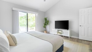 Ein Schlafzimmer mit einem Bett, einem Fernseher auf einer weißen Kommode, einer Pflanze und einer Glastür zu einem Balkon.