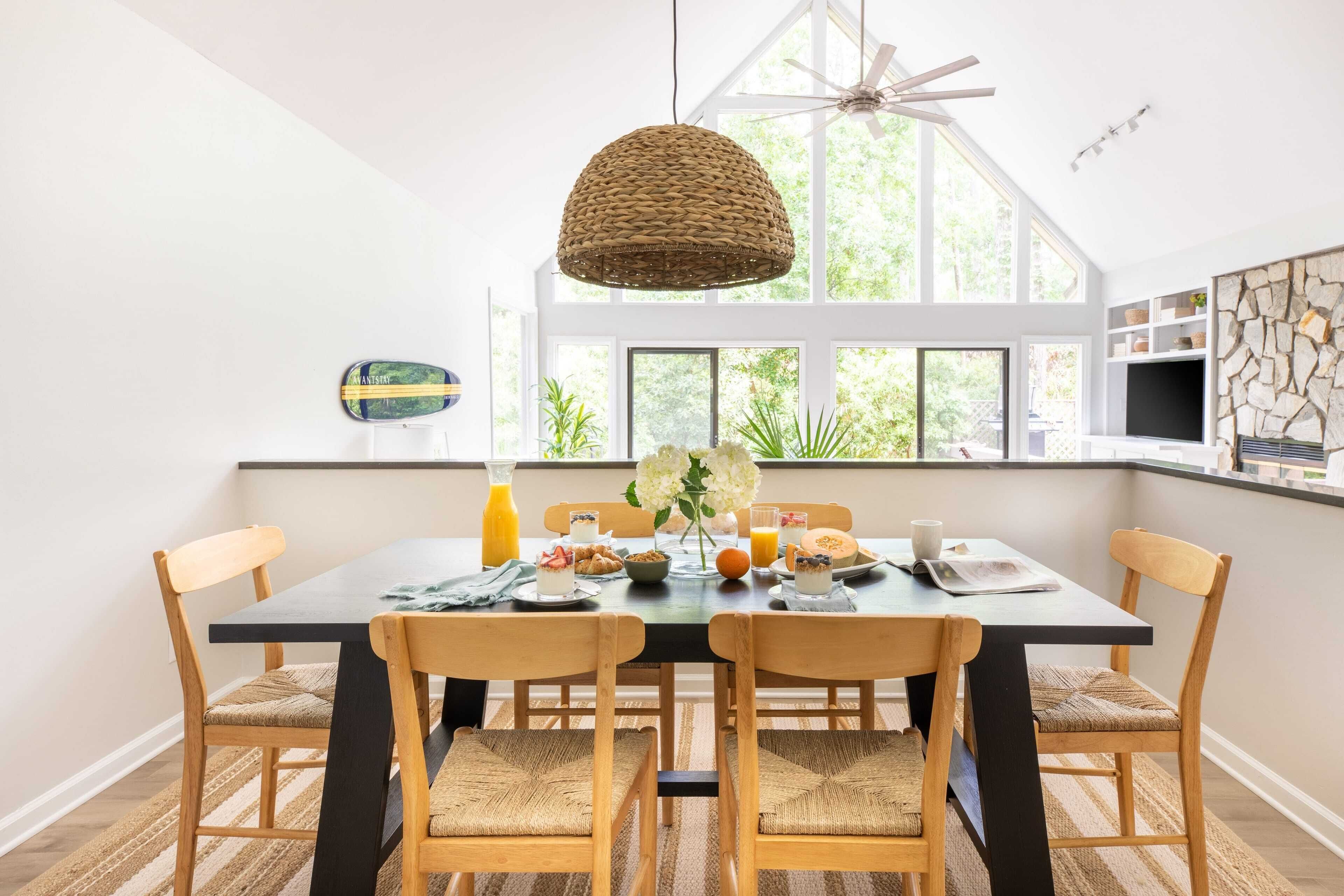 A dining area with a large table, four chairs, a woven pendant light, and a view of greenery through large windows.