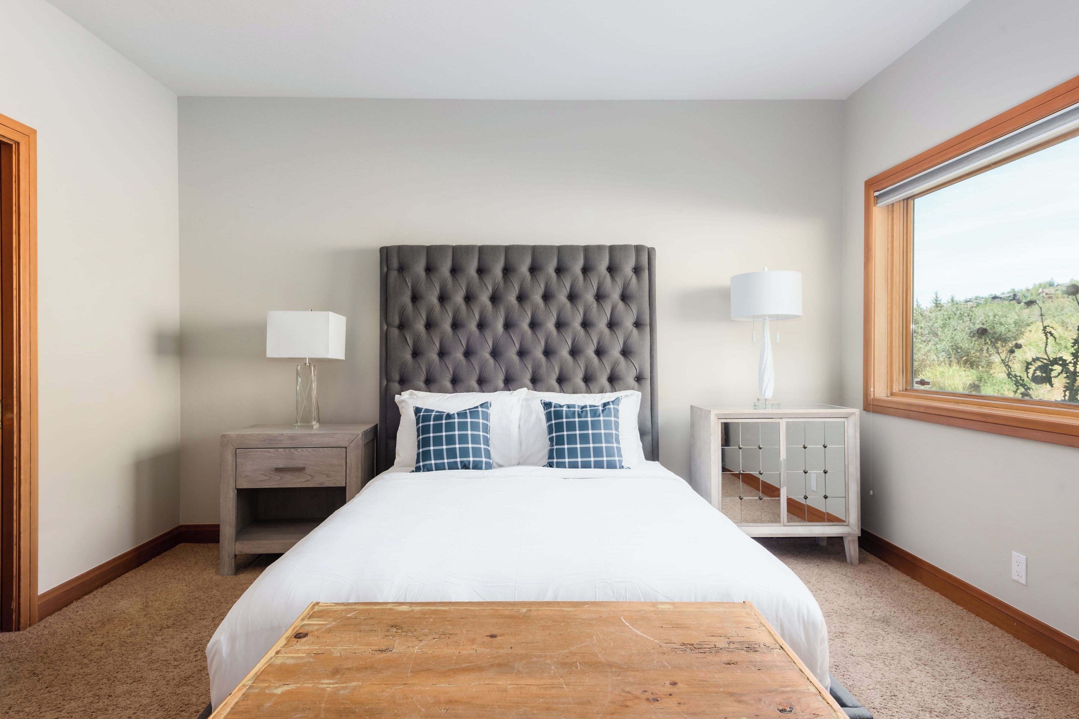 Une chambre à coucher avec une tête de lit capitonnée, une table de chevet en bois, une table d’appoint, une fenêtre donnant sur un beau paysage et une table en bois placée au centre de la pièce.