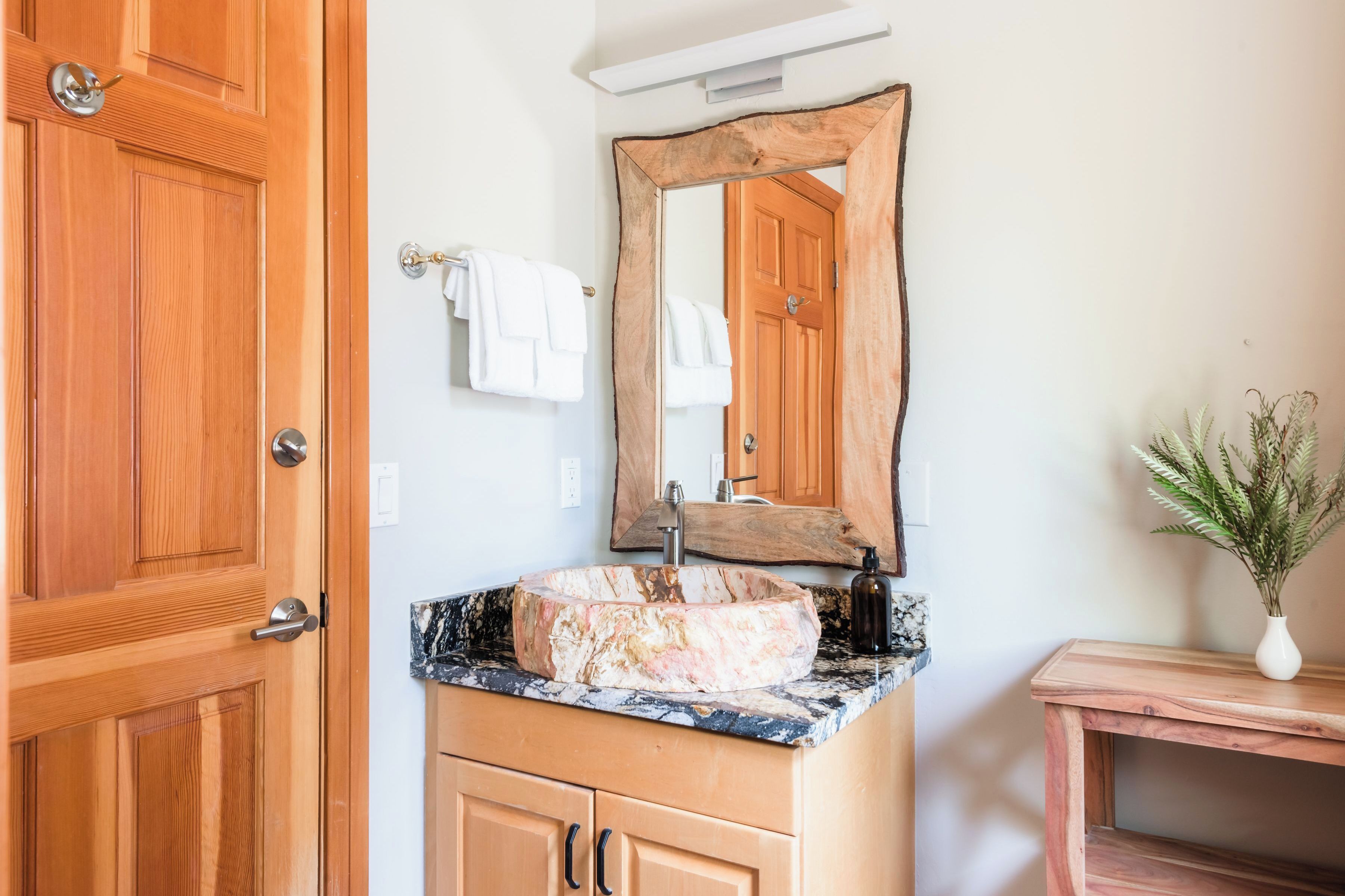 A bathroom with a wooden door, a marble sink, a wooden vanity, a mirror with a wooden frame, and a potted plant.