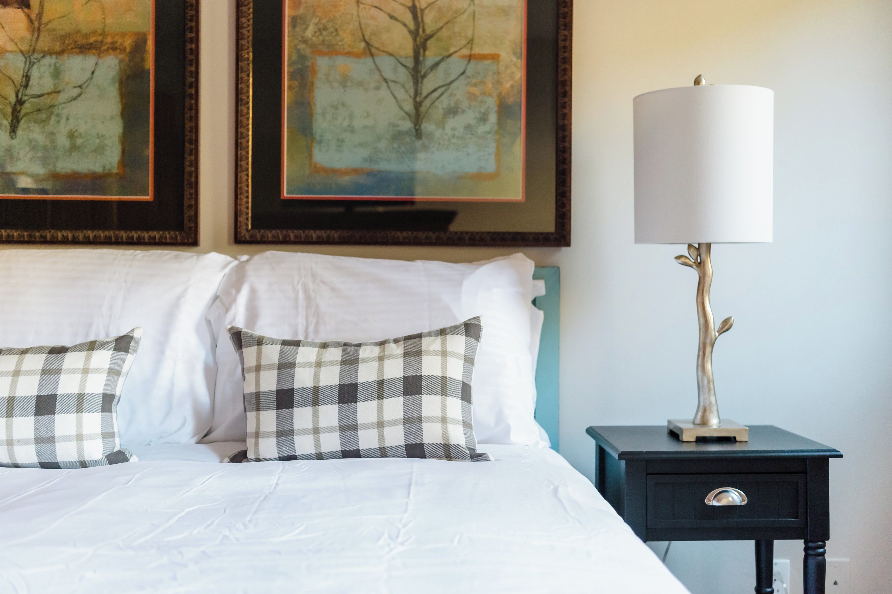A neatly made bed with white linens and checkered pillows, a bedside table with a lamp, and two framed artworks on the wall.