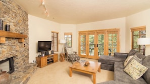 A living room with a stone wall, a fireplace, a television, a sofa, and a dining table. - Westermere 311 - 2 by Avantstay Just Steps From Ski Area w/ Great Views! (Telluride)