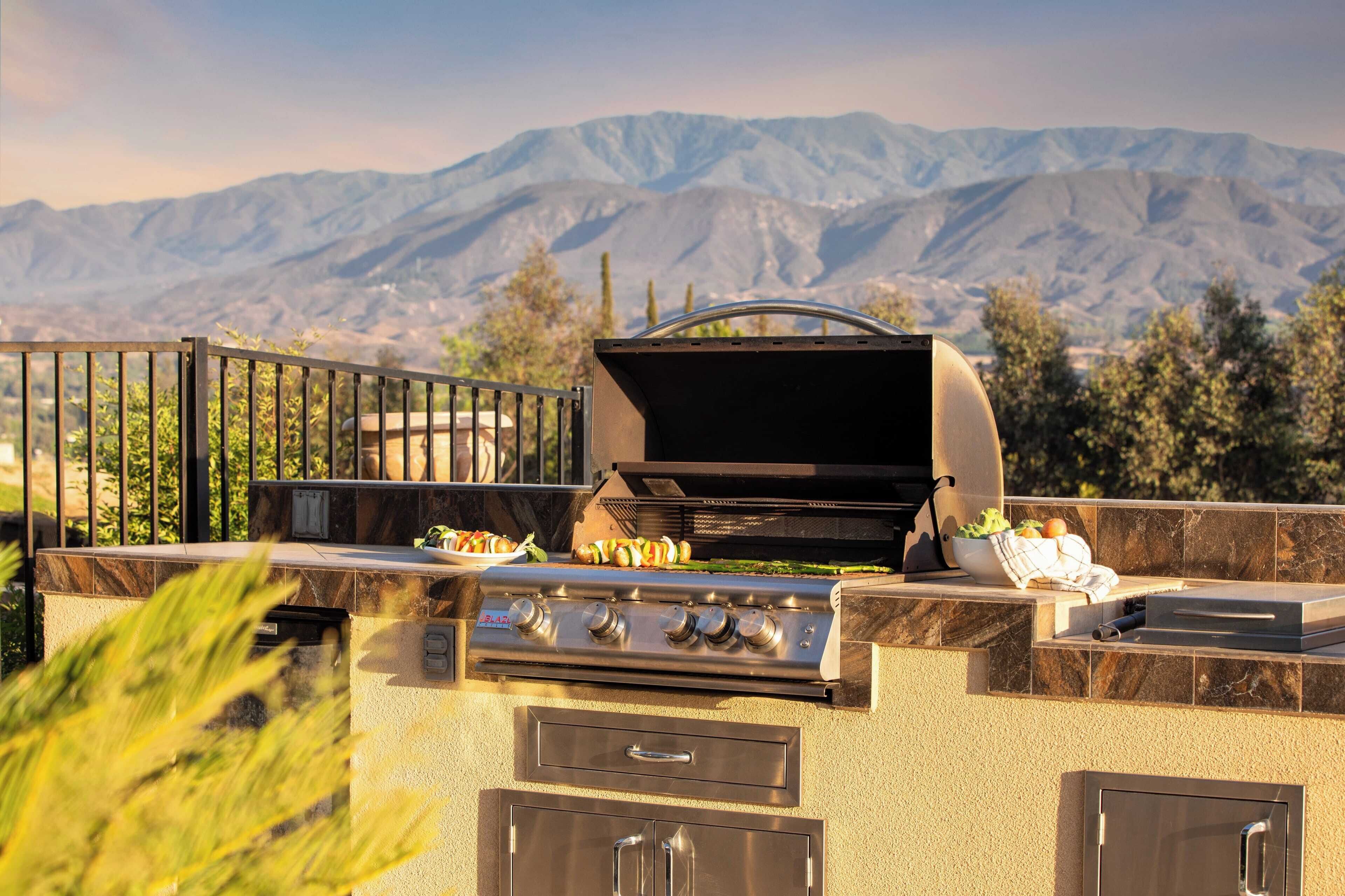 Une cuisine extérieure équipée d’un barbecue et d’une cuisinière, entourée d’un mur en pierre et d’une clôture en métal, avec des montagnes en arrière-plan.