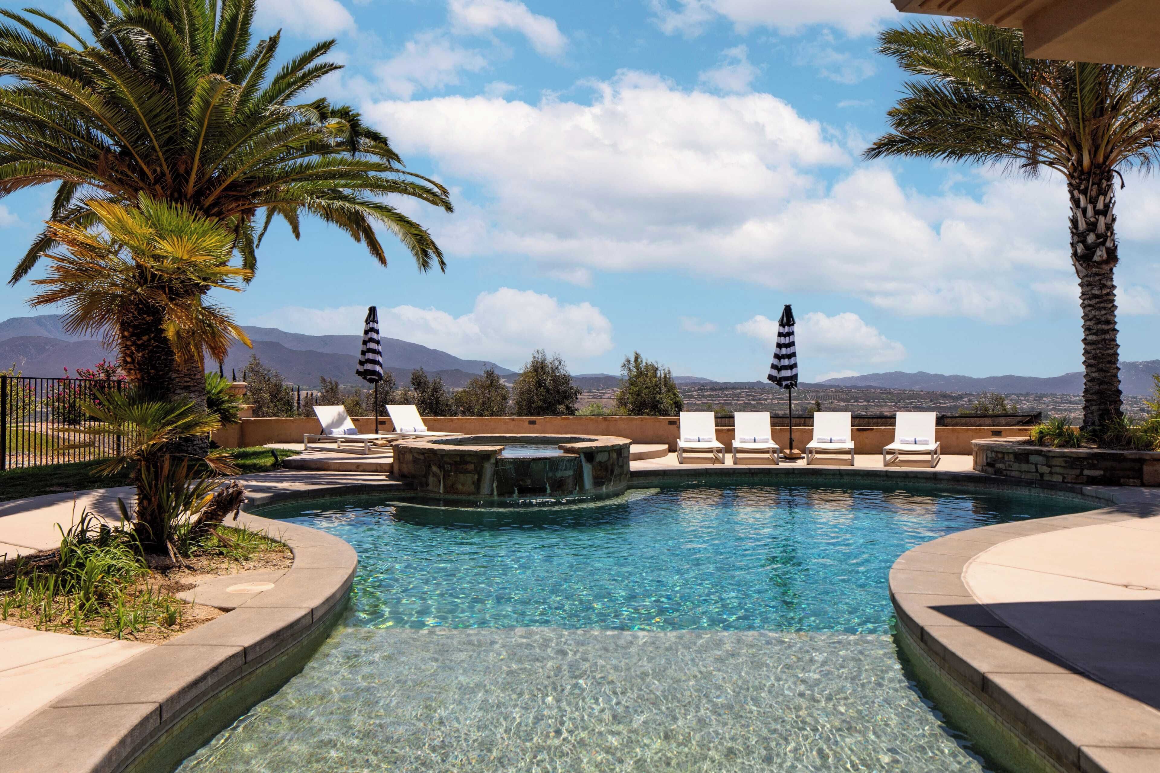 A clear swimming pool with lounge chairs and palm trees, surrounded by mountains.