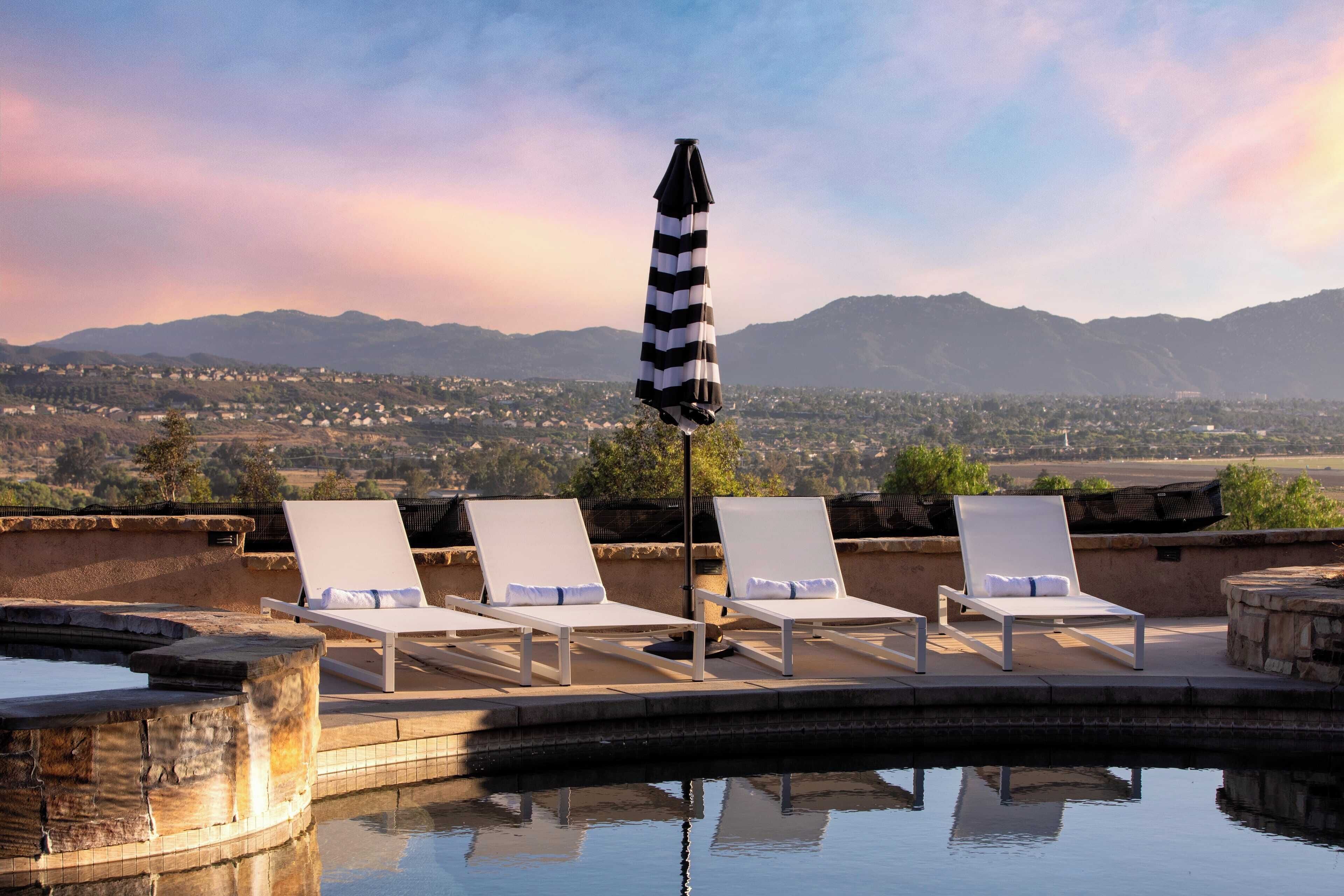A poolside area with lounge chairs and a striped umbrella, overlooking a mountainous landscape.