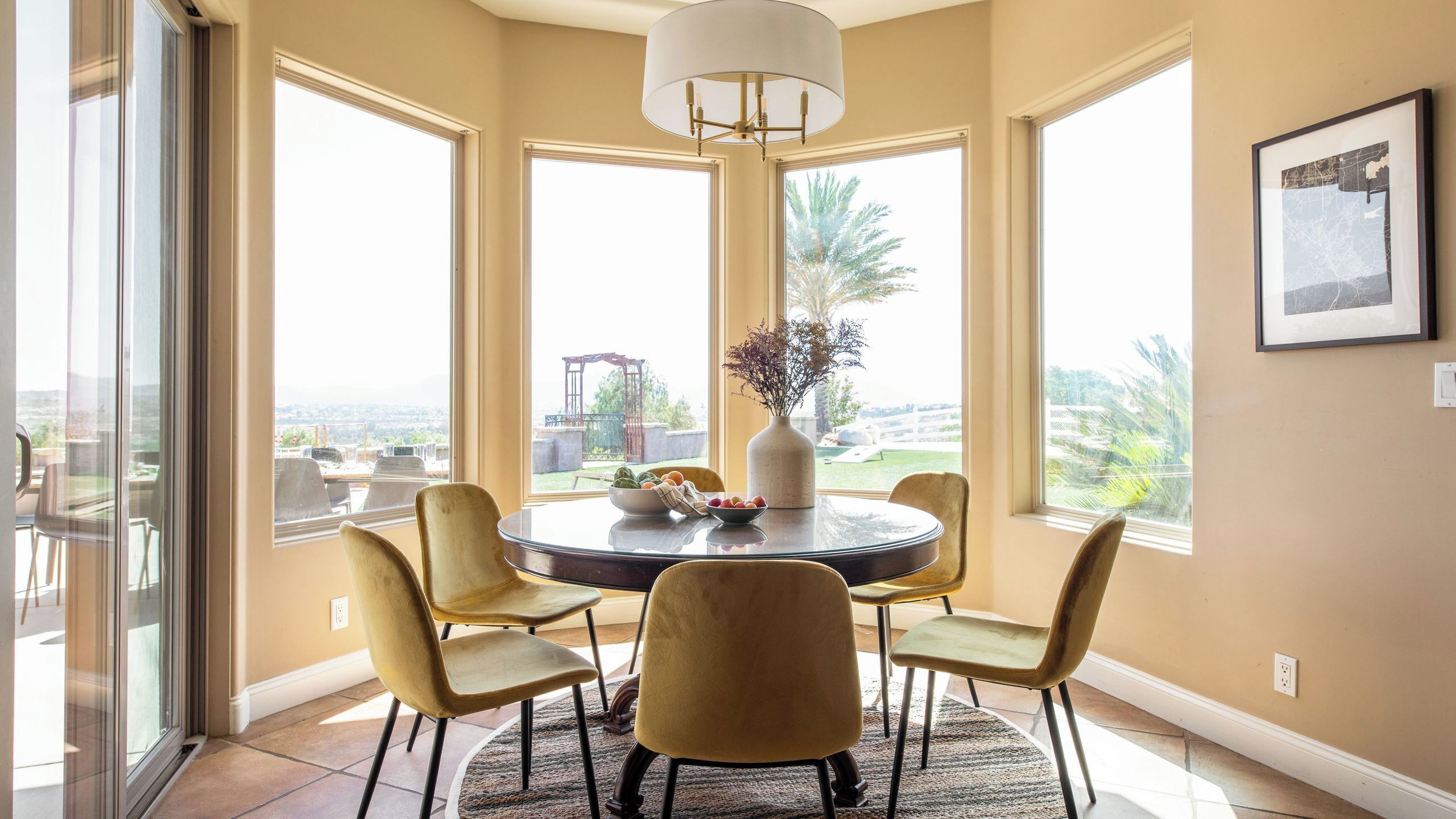 A dining area with a round table, four chairs, a vase with flowers, and a view of the outdoors.