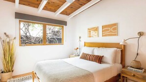 Un dormitorio con una cama grande, una ventana con vistas a árboles, una mesita de noche con lámpara y dos obras de arte en las paredes.