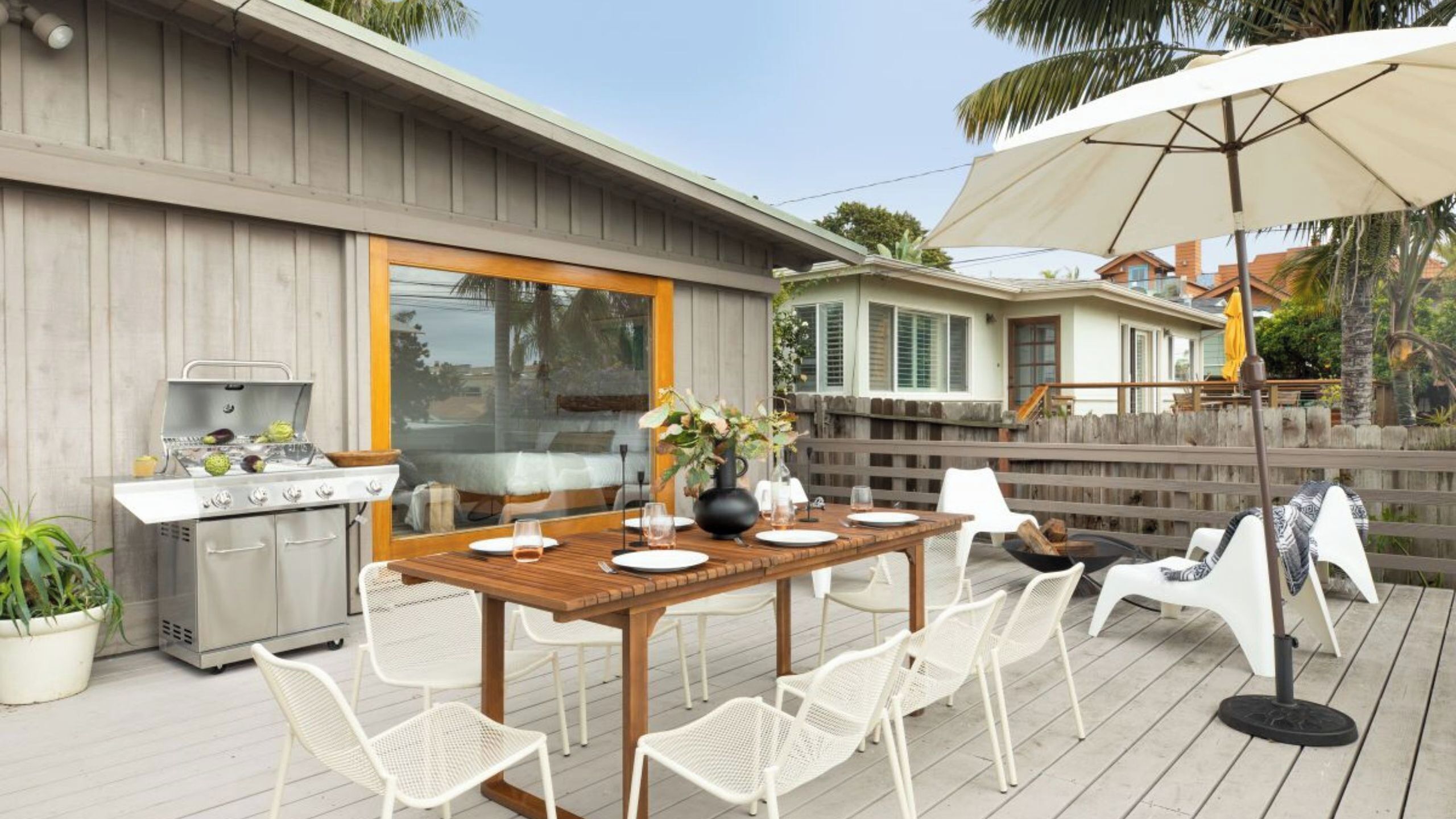 An outdoor patio with a wooden table, white chairs, a grill, and a large umbrella.